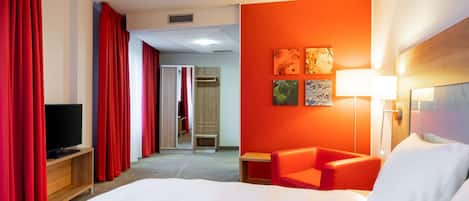 A hotel room with a large bed, red curtains, a TV on a wooden stand, a red armchair, and a wall with three framed artworks.