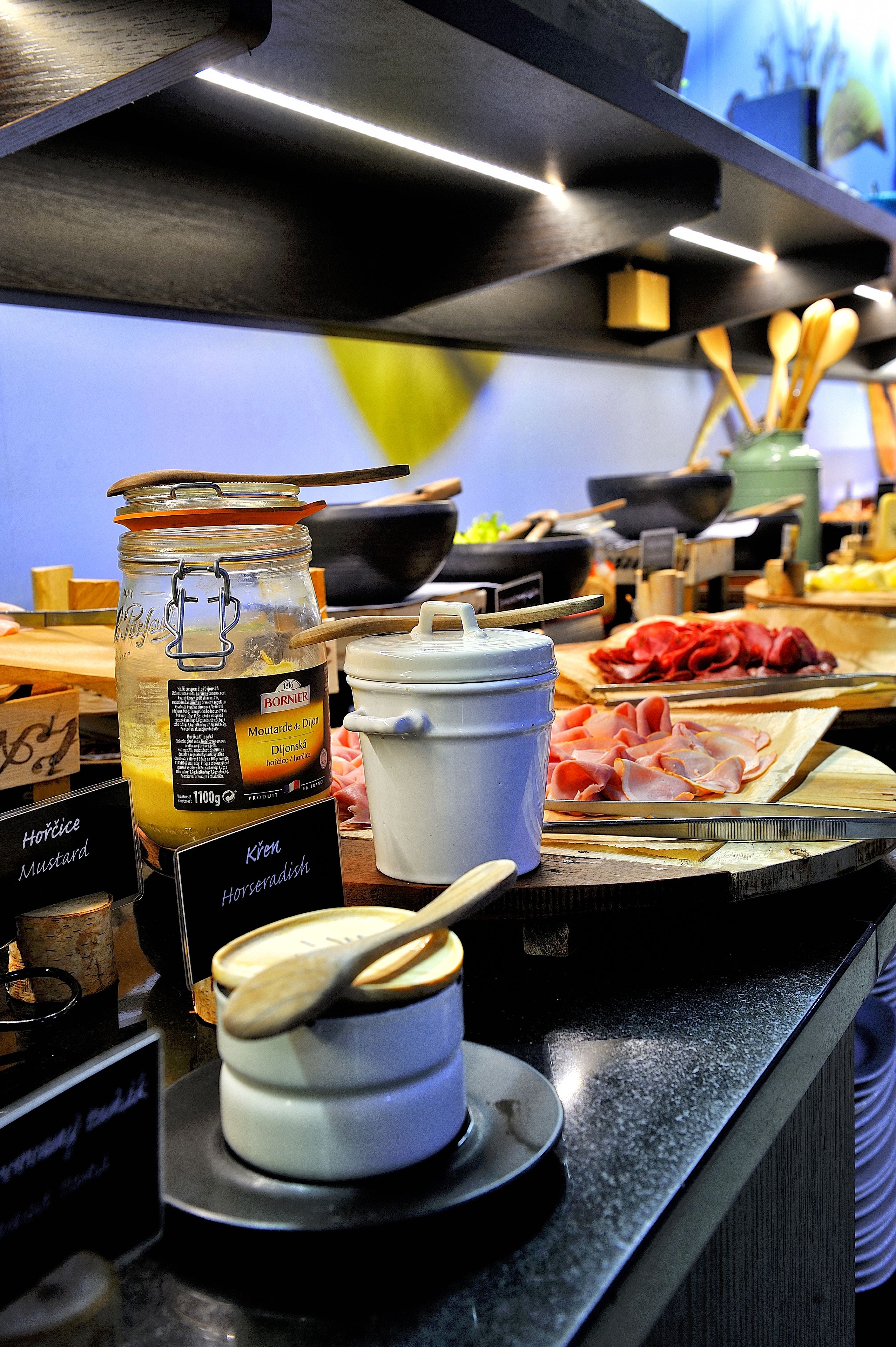 Un buffet proposant une variété de plats, notamment un pot de moutarde, un récipient blanc doté d’un couvercle et plusieurs ustensiles en bois.
