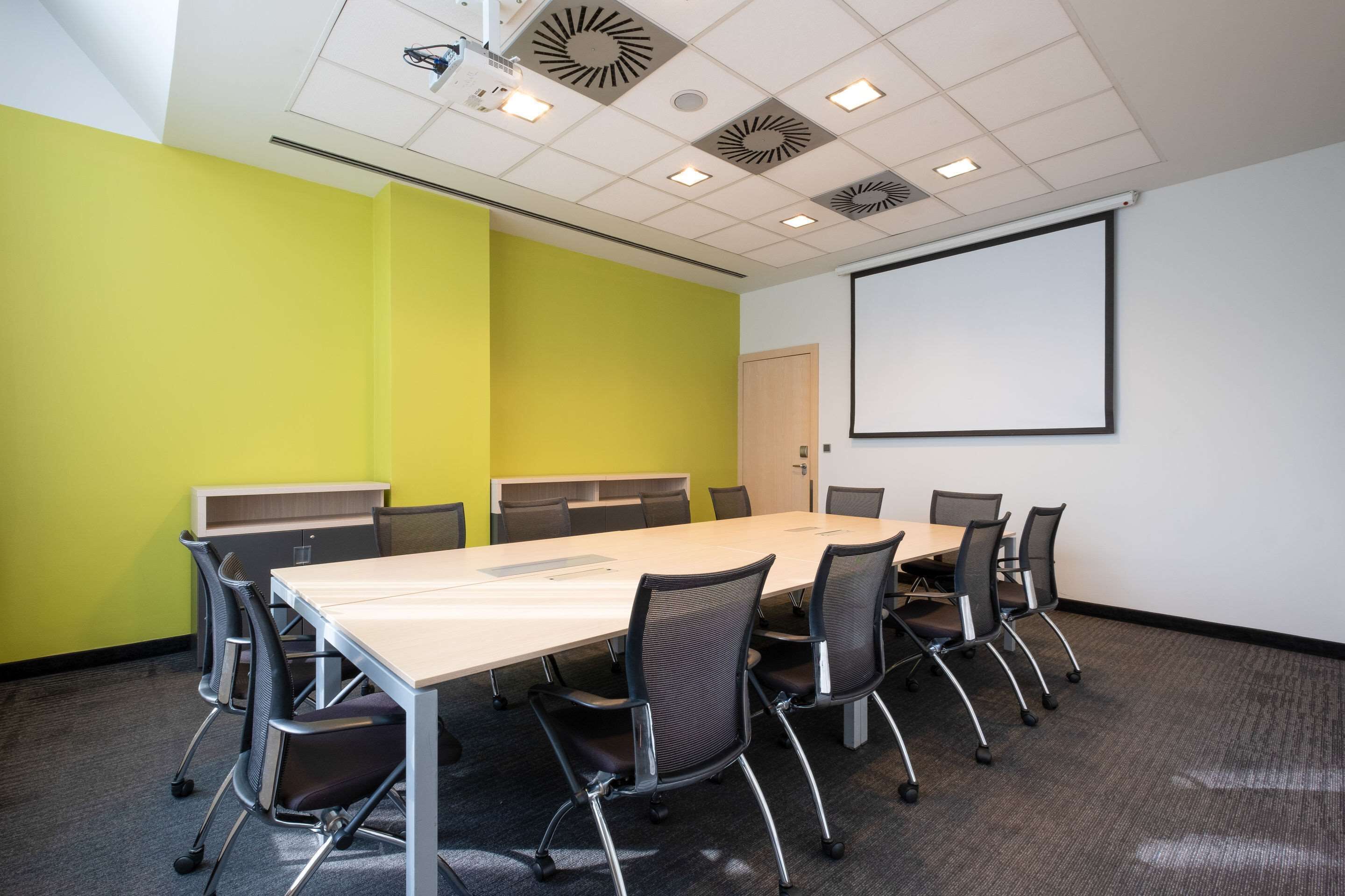 Una sala de conferencias con una larga mesa, sillas, una pantalla de proyector y una pizarra.
