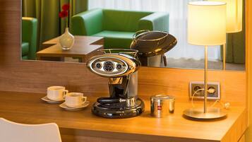 A hotel room with a coffee machine, two cups, a vase with flowers, a lamp, and a mirror reflecting a green couch and curtains.