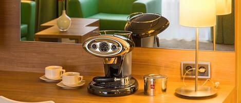 A hotel room with a coffee machine, two cups, a vase with flowers, a lamp, and a mirror reflecting a green couch and curtains.