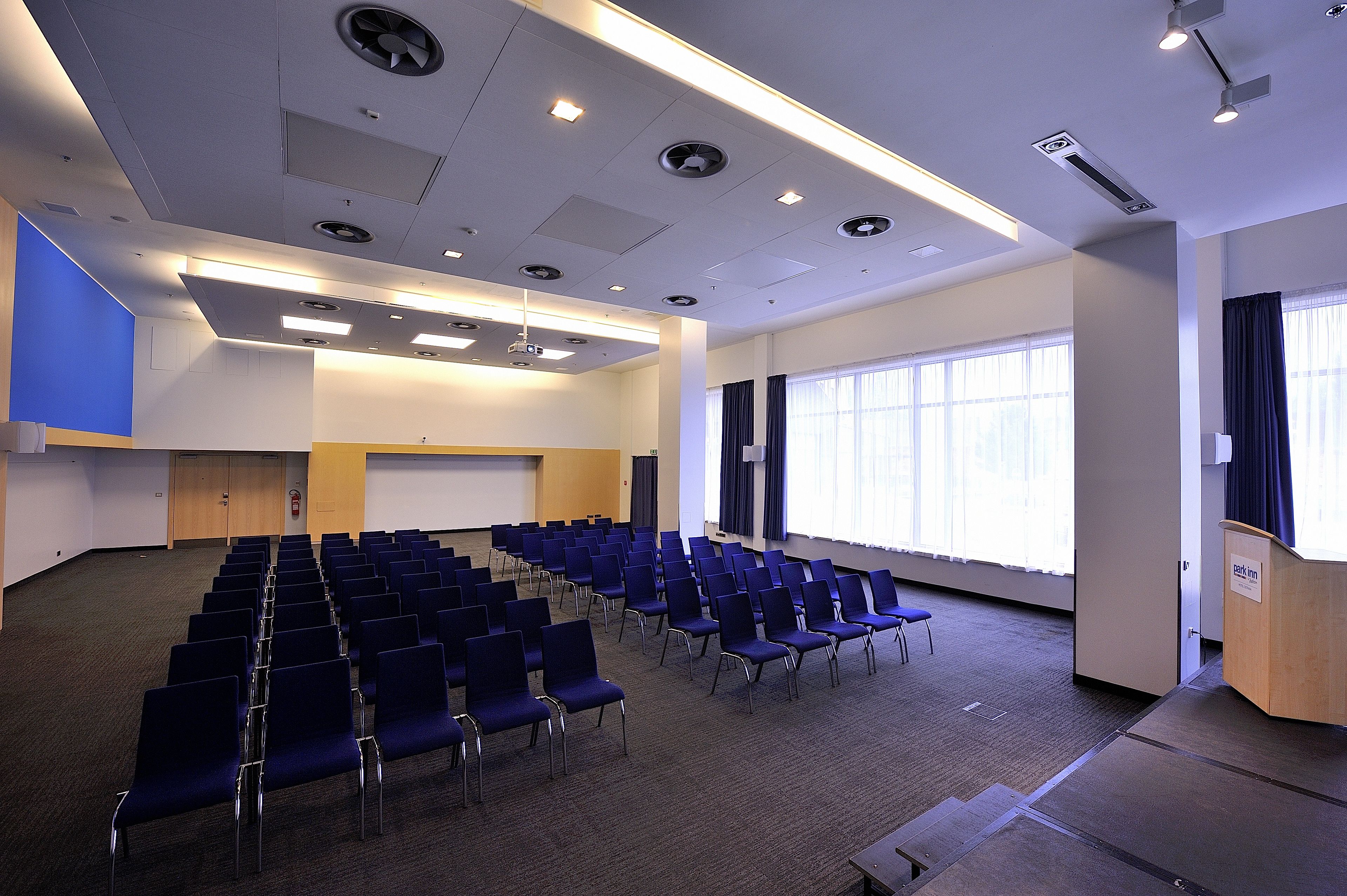 Un salón de conferencias con filas de sillas, un atril y una pantalla para proyector.