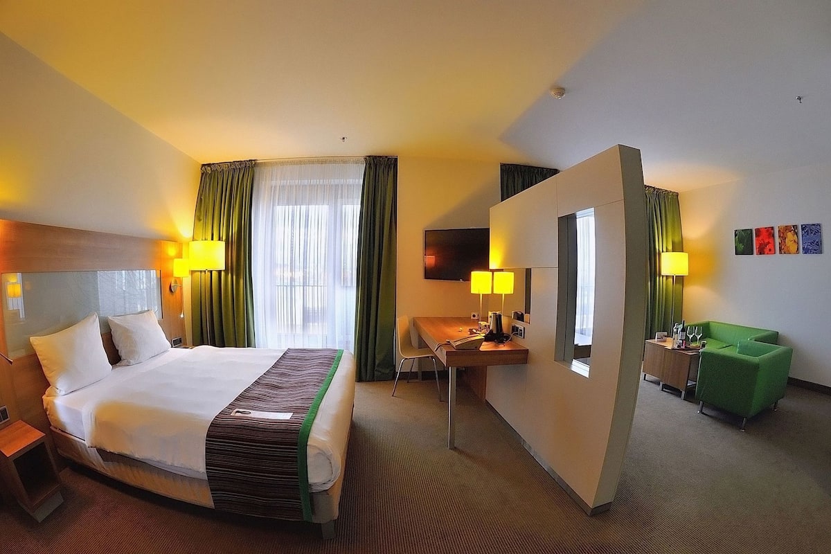 Ein Hotelzimmer mit Bett, Schreibtisch, Stuhl und einem grünen Sessel.