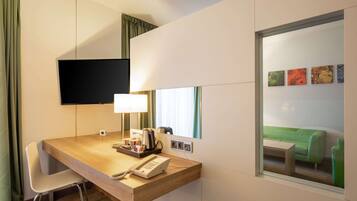 A hotel room with a wooden desk, a telephone, a lamp, a flat-screen TV mounted on the wall, and a large window with a green couch visible through it.