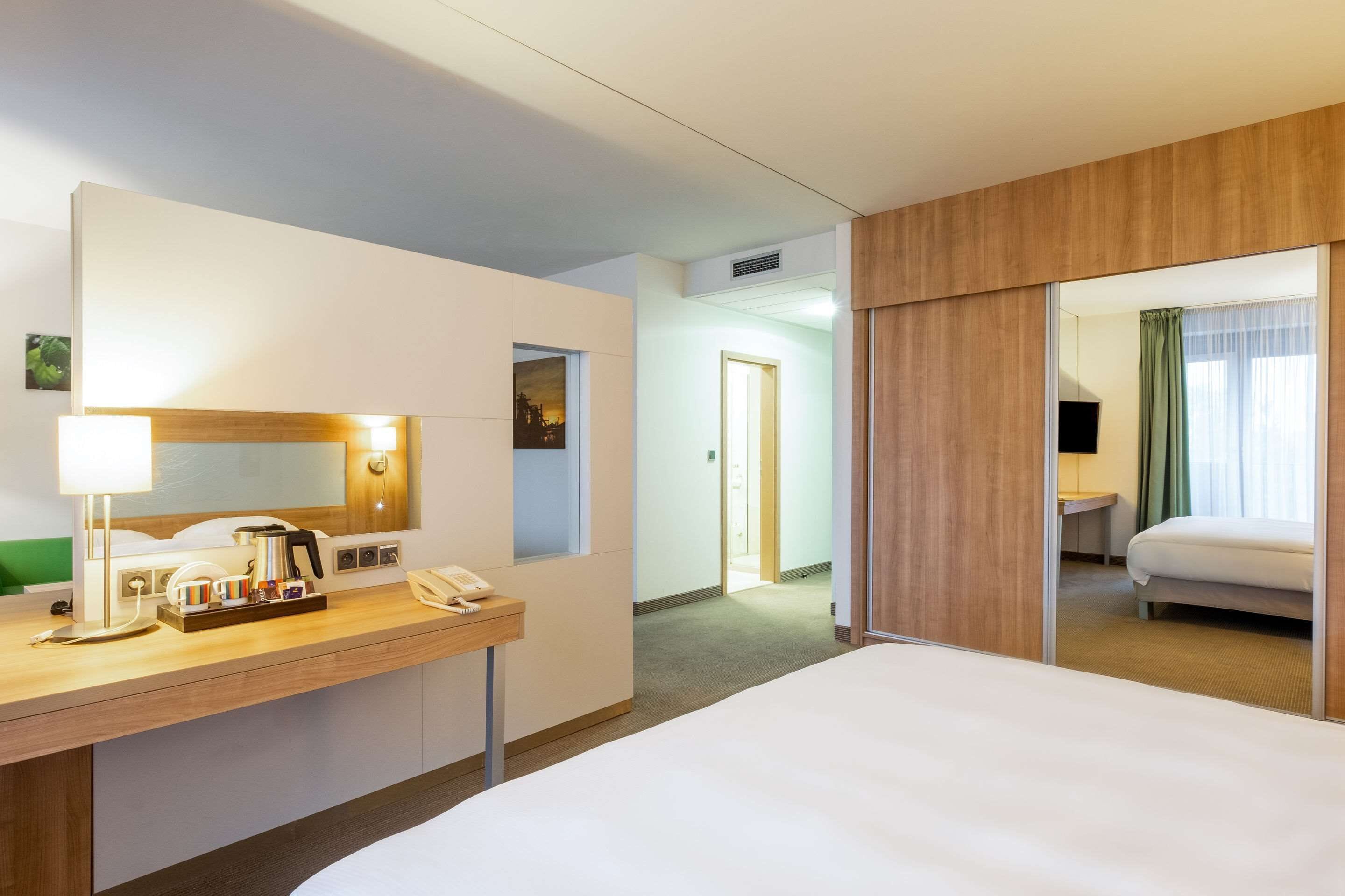 Une chambre d’hôtel avec un lit, un bureau doté d’une lampe, un téléphone et un miroir. Une porte donne accès à une autre pièce.