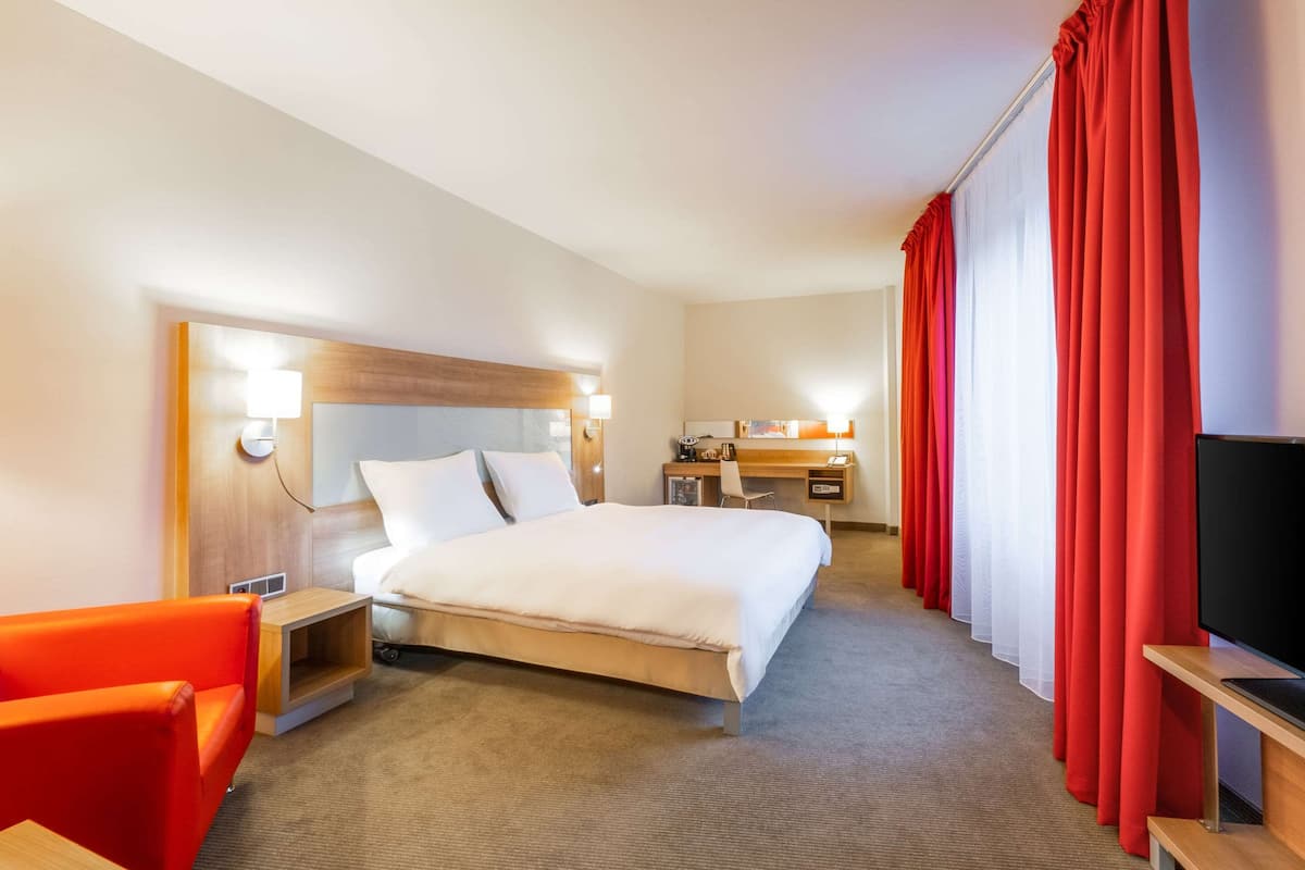 Ein Hotelzimmer mit einem Bett, einem Schreibtisch, einem Sessel, einem Fernseher und roten Vorhängen.