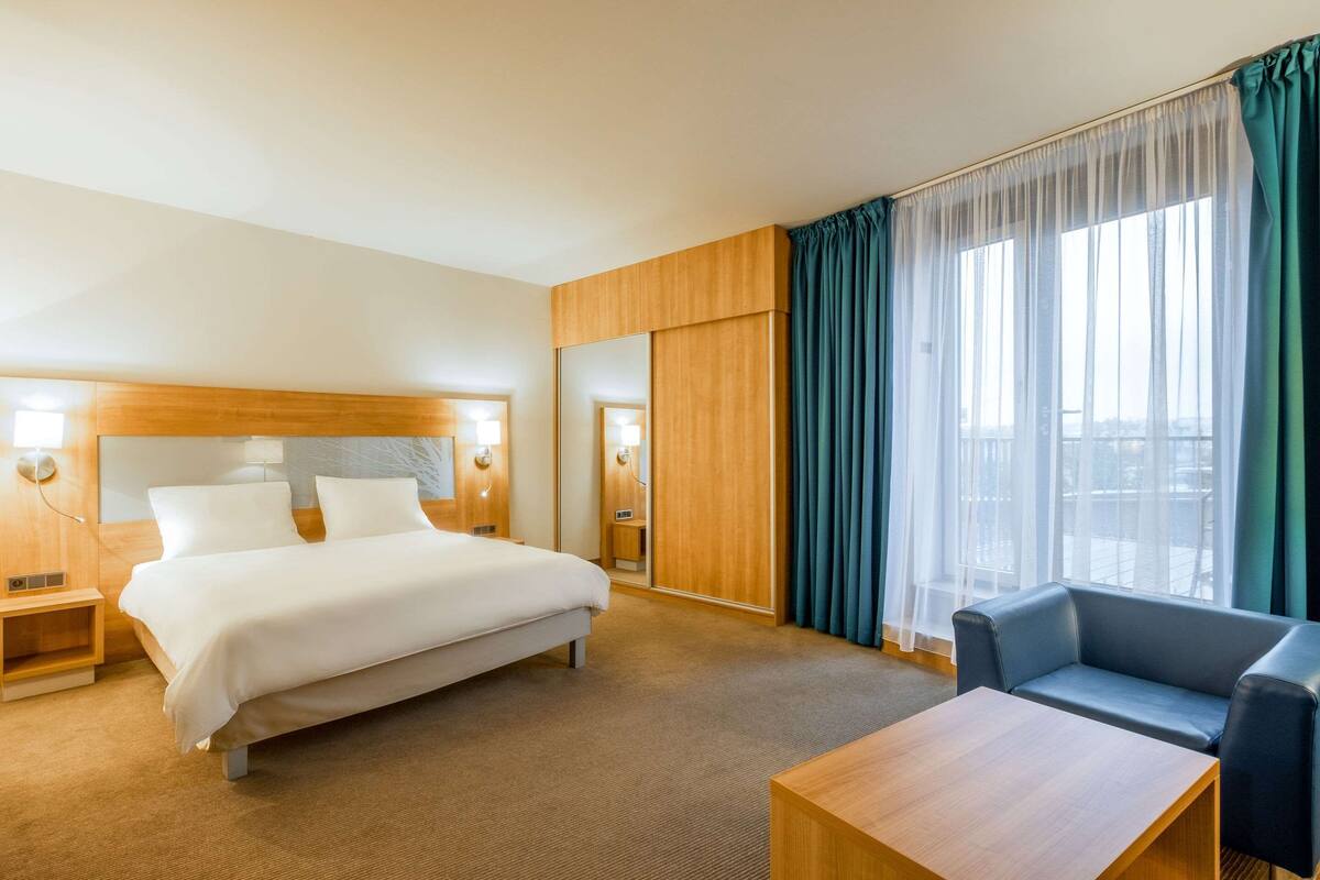 Ein Hotelzimmer mit einem großen Bett, einem Holzkopfteil, einem Nachttisch aus Holz, einem Schreibtisch, einem Stuhl und einem großen Fenster mit Vorhängen.