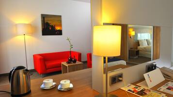 Habitación de hotel con sofá rojo, una estación de café, lámpara de noche, espejo y una pintura de paisaje urbano en la pared.