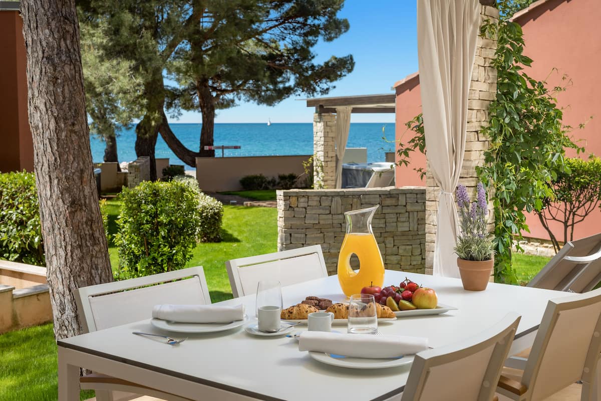 A table set for breakfast with a view of the sea, outdoor seating, and a potted plant.