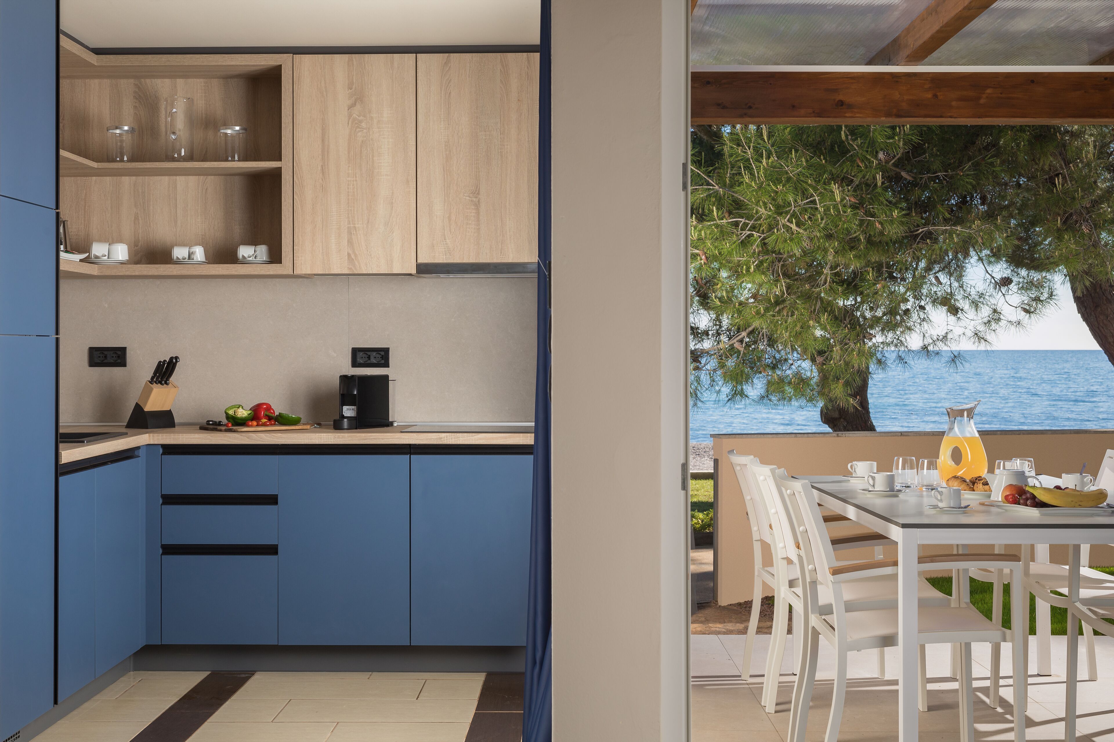 Una cocina moderna con armarios azules, una cafetera y un frutero. Al exterior, hay un comedor con una mesa y sillas, con vista a un cuerpo de agua.