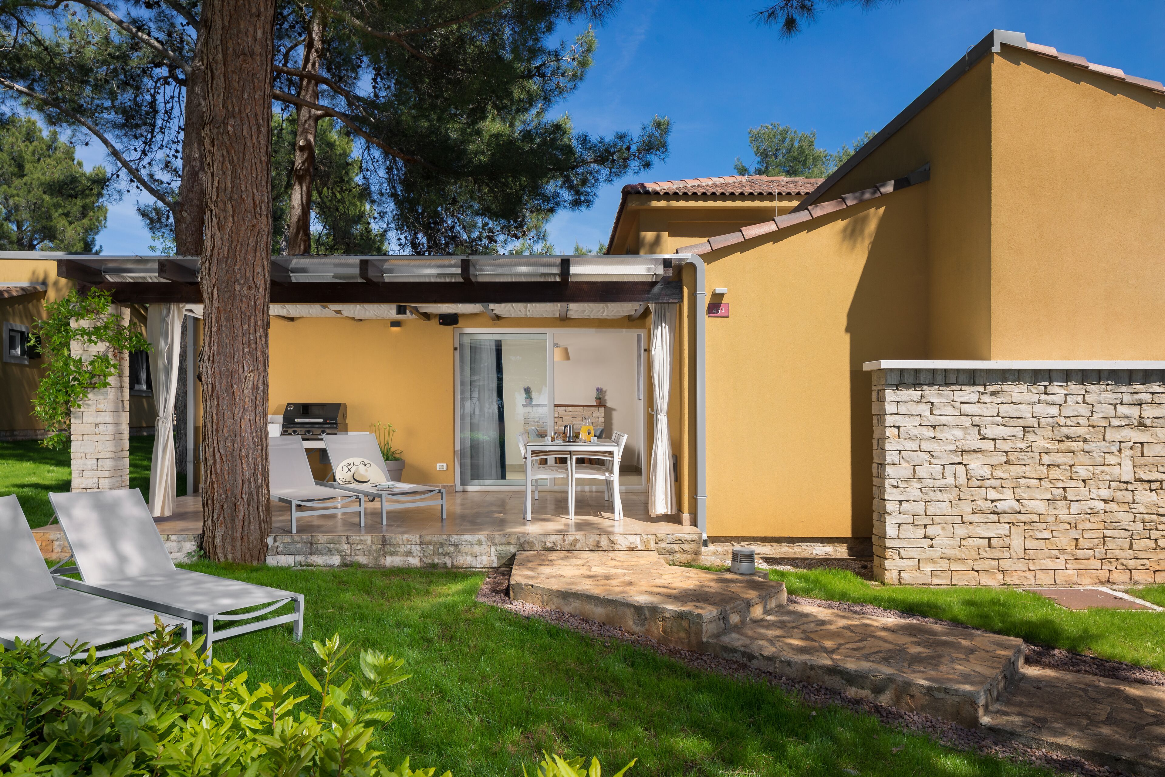 Un edificio giallo con un muro in pietra, un portico coperto con poltrone relax e un’area barbecue.