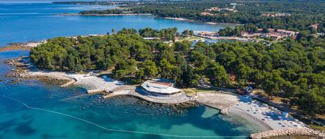 Vue aérienne d’une zone côtière avec une plage, une petite île abritant des bâtiments et une mer d’un bleu limpide.