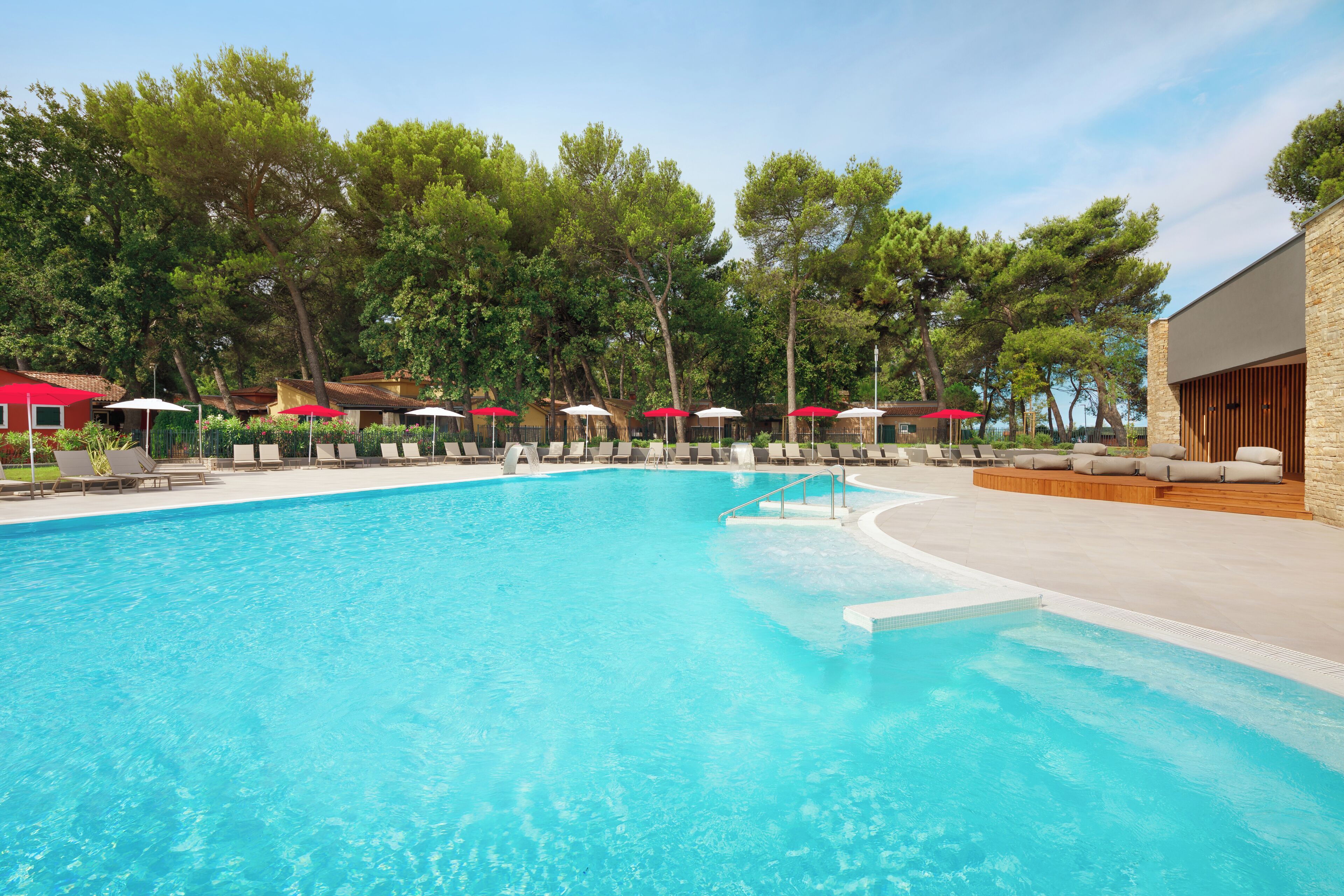 A swimming pool with lounge chairs and umbrellas, surrounded by trees and a building with a stone facade.