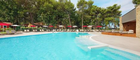 A swimming pool with lounge chairs and umbrellas, surrounded by trees and a building with a stone facade.
