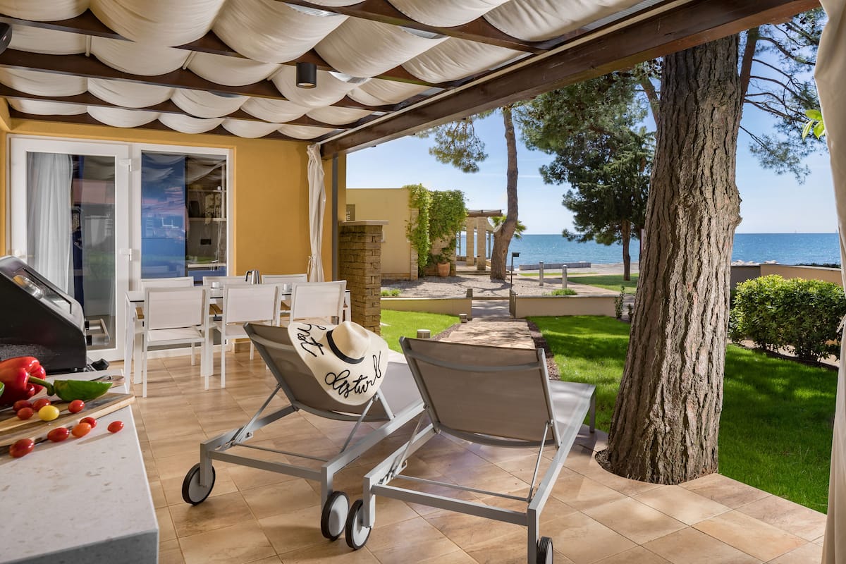 A covered outdoor seating area with a grill, chairs, and a view of the sea.