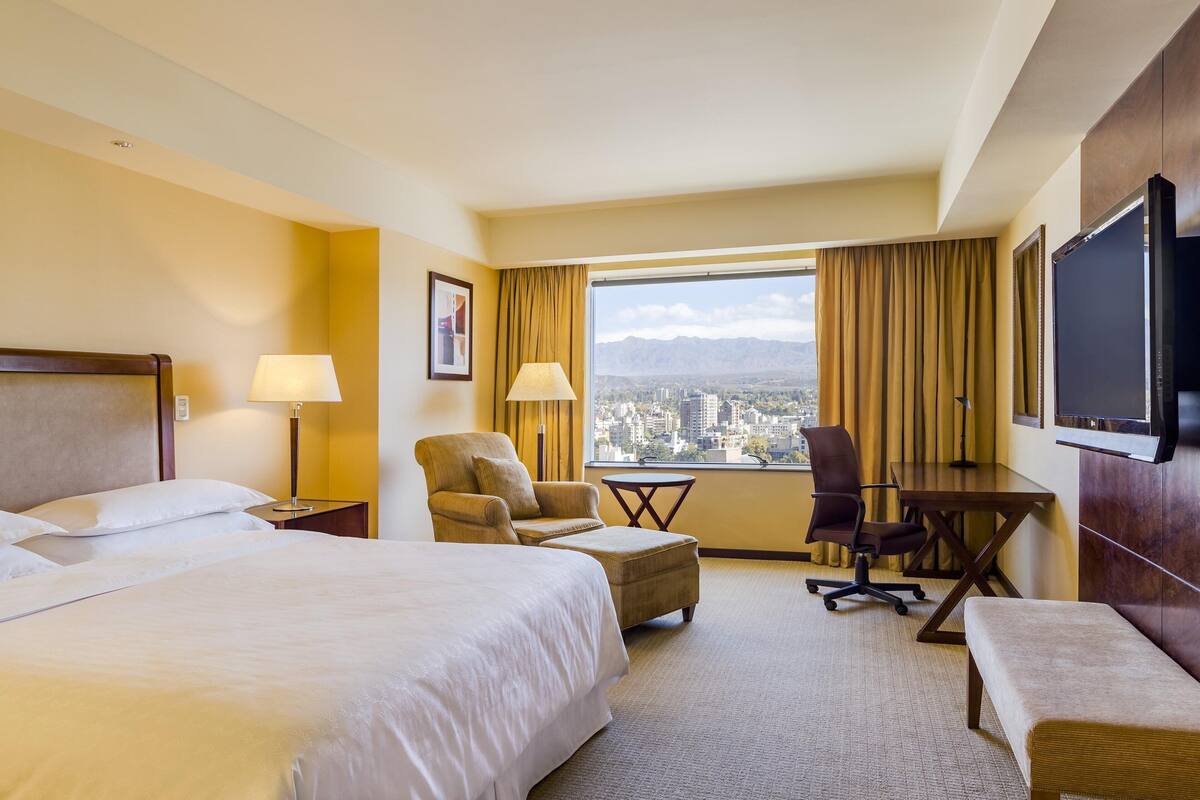 Une chambre d’hôtel avec un grand lit, un bureau, une chaise, une petite table, un téléviseur et une vue sur le paysage urbain, visible à travers une grande fenêtre.
