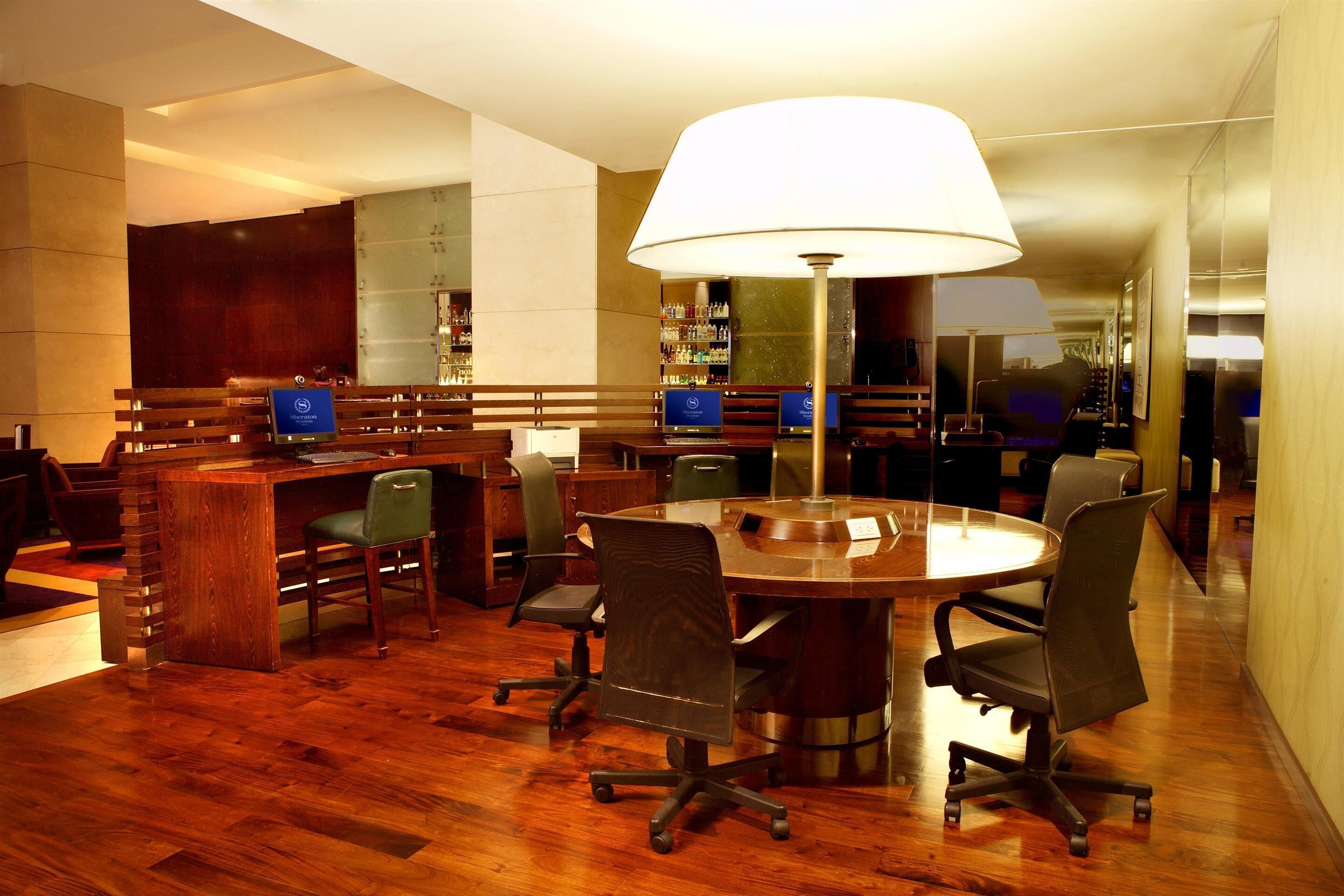A conference room with a round table, several chairs, a desk with a computer, and a large lamp.