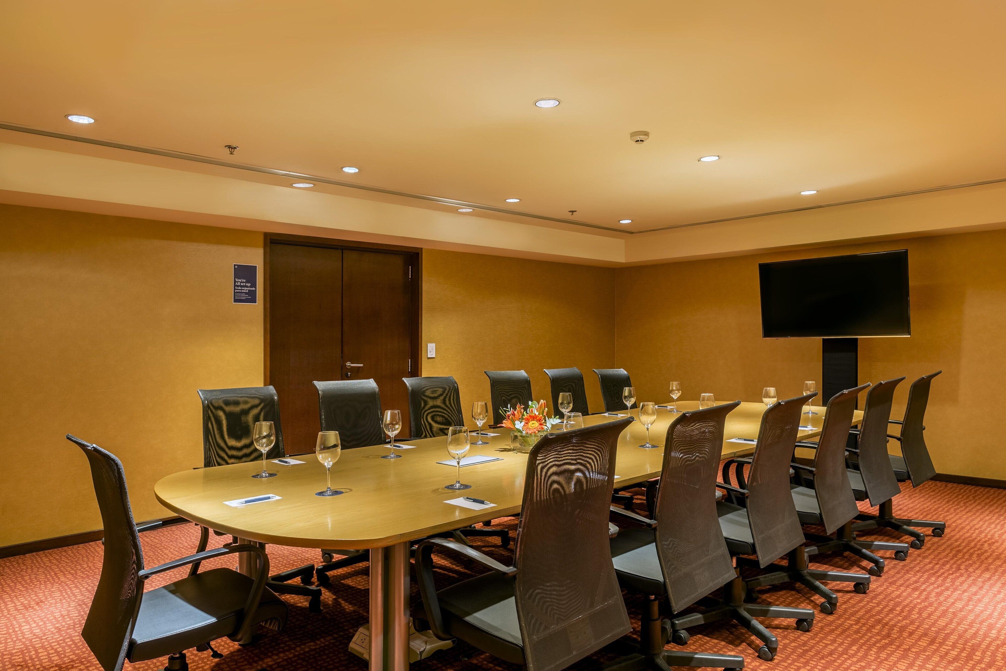 Uma sala de conferências com uma mesa comprida, cadeiras e uma televisão de tela plana na parede.