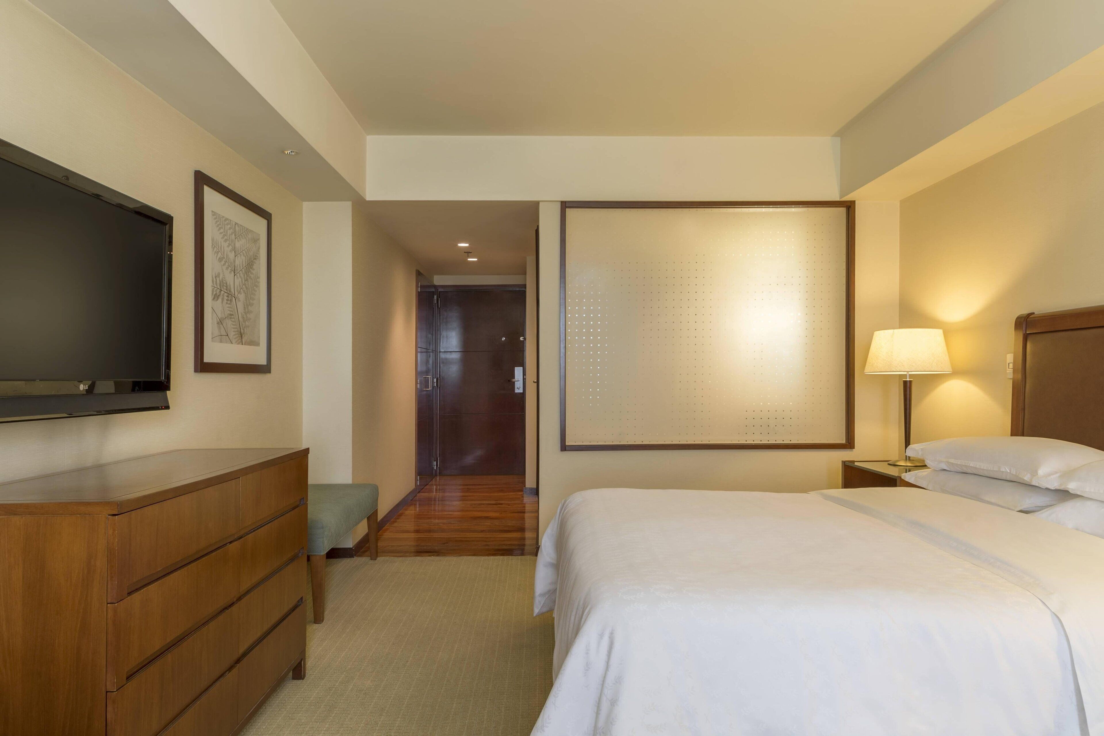 A hotel room with a bed, a dresser, a television, a wall-mounted artwork, a door, and a nightstand with a lamp.