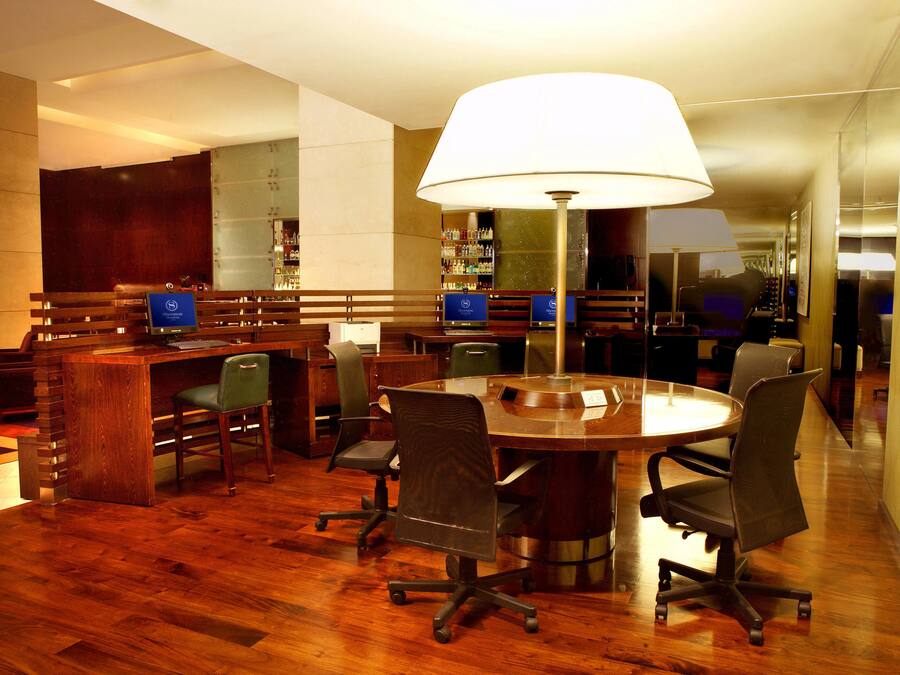 Uma sala de conferências com uma mesa redonda, quatro cadeiras, uma escrivaninha e um abajur grande.