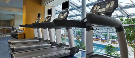 A row of treadmills in a modern indoor gym with a glass ceiling and a view of an outdoor pool area.