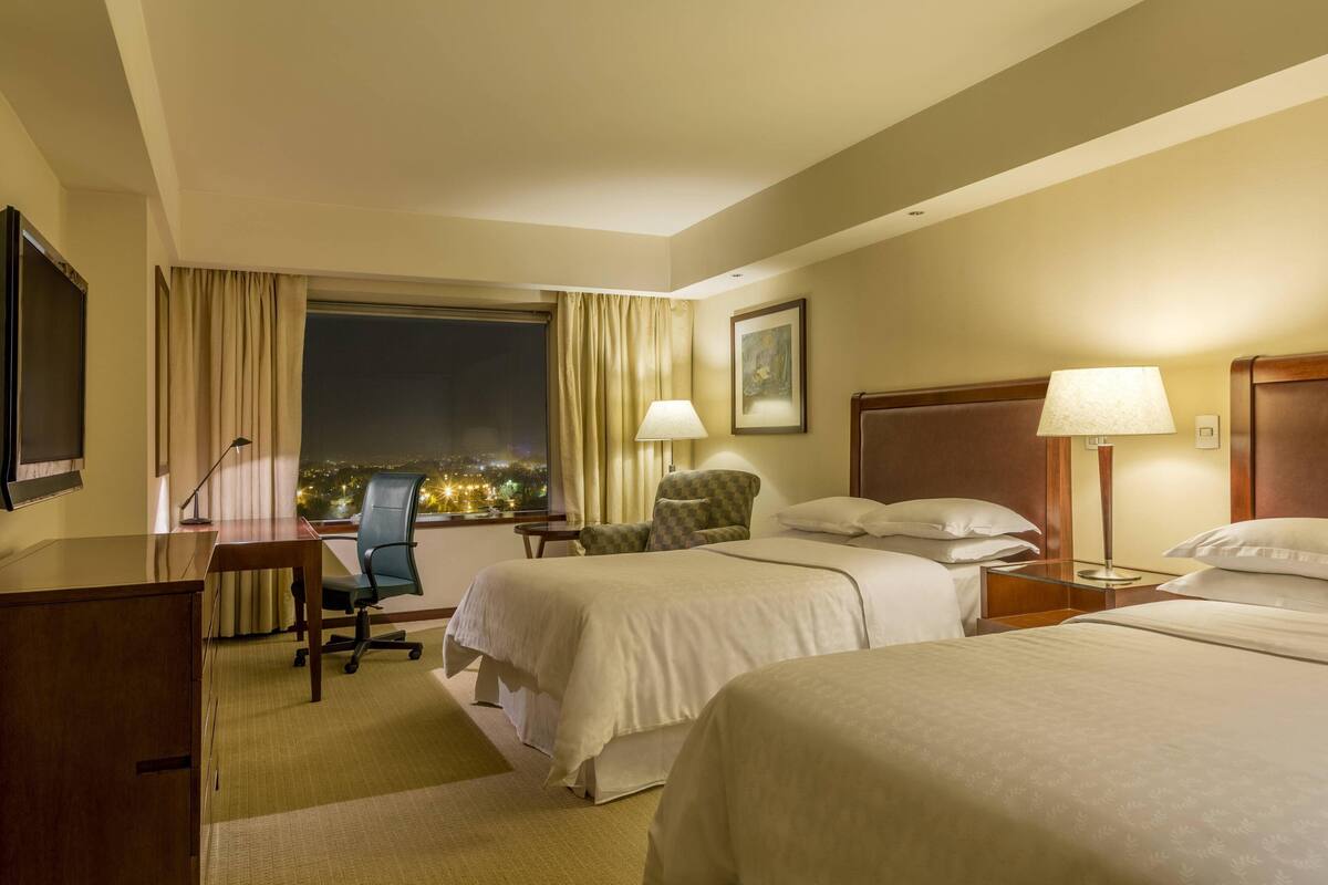 Une chambre d’hôtel avec deux lits, un bureau, une chaise, une lampe et une fenêtre donnant sur la ville.