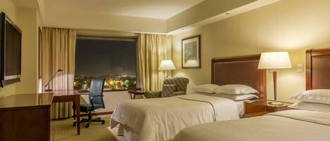 Habitación de hotel con dos camas, un escritorio, una silla, una lámpara y ventana con vista a la ciudad.