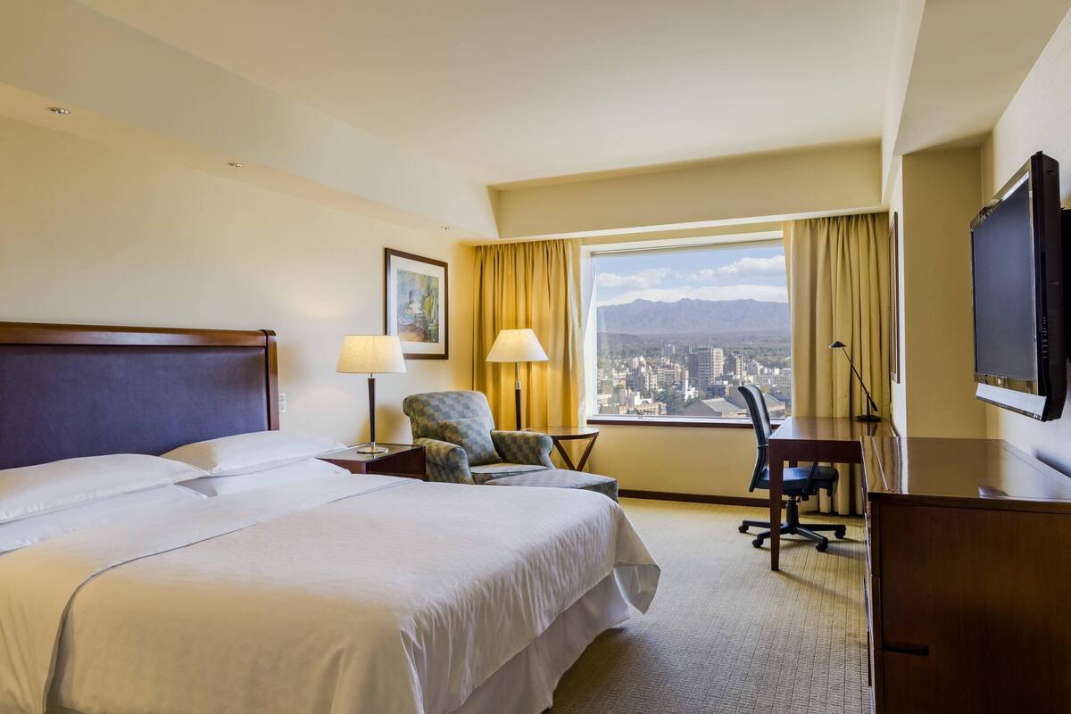 Une chambre d’hôtel avec un grand lit, un bureau, une chaise, une télévision et une fenêtre donnant sur la ville.