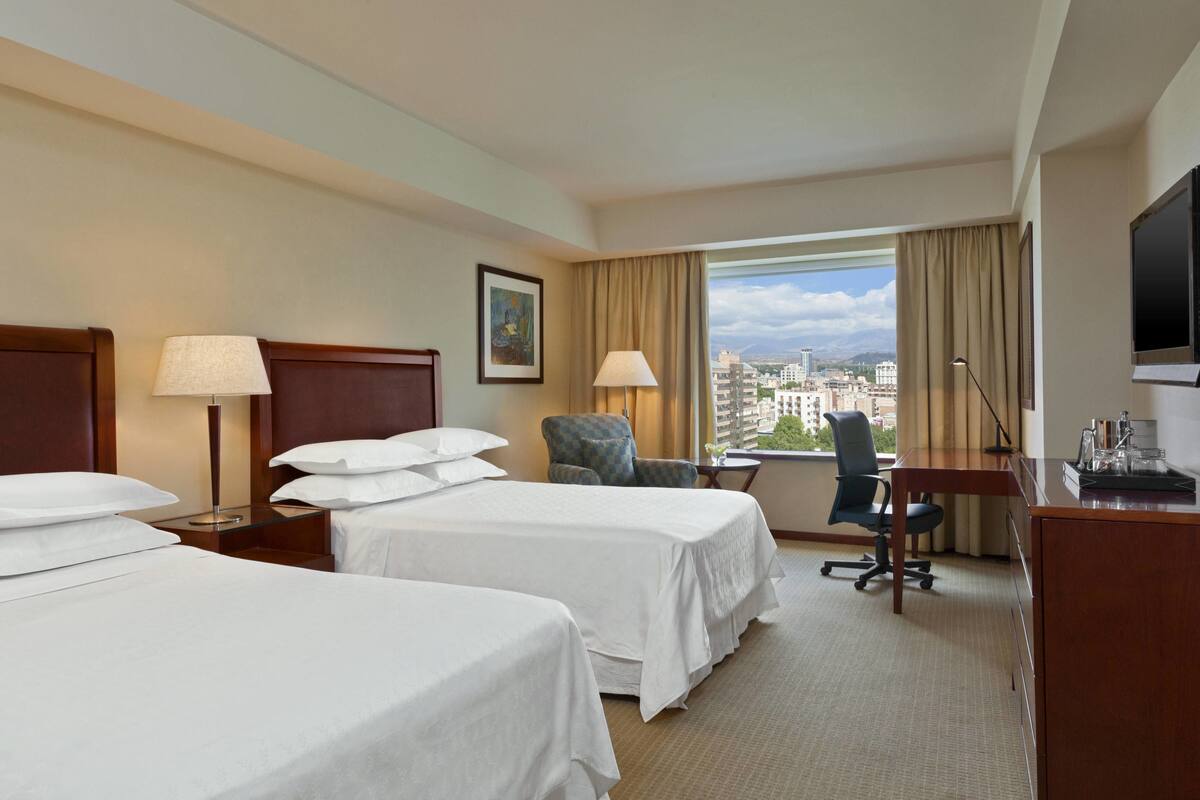 Une chambre d’hôtel avec deux lits, un bureau, une chaise, une télévision et une fenêtre donnant sur la ville.