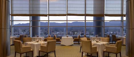 A dining area with tables set for six, large windows with a cityscape view, and a high ceiling.