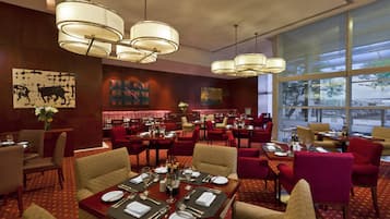 A restaurant with multiple dining tables, red chairs, and hanging light fixtures.