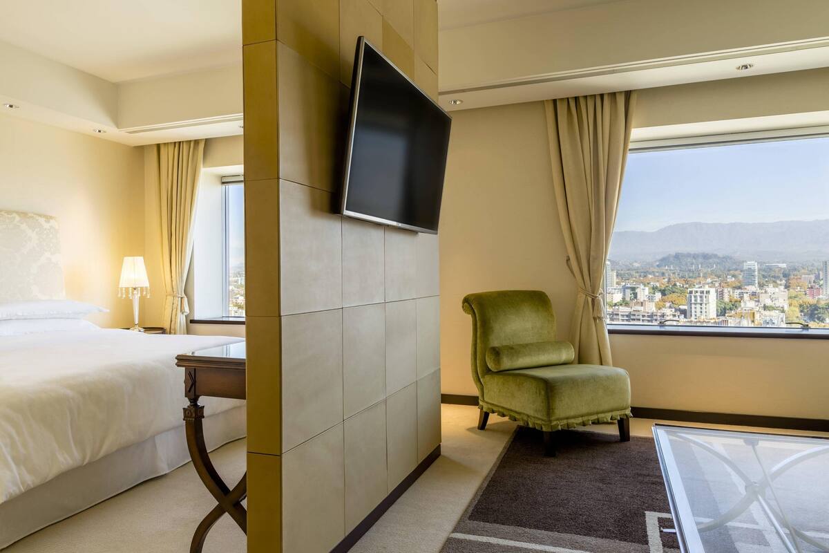 Une chambre d’hôtel avec un lit, un téléviseur fixé au mur, une chaise et une vue sur le paysage urbain par la fenêtre.
