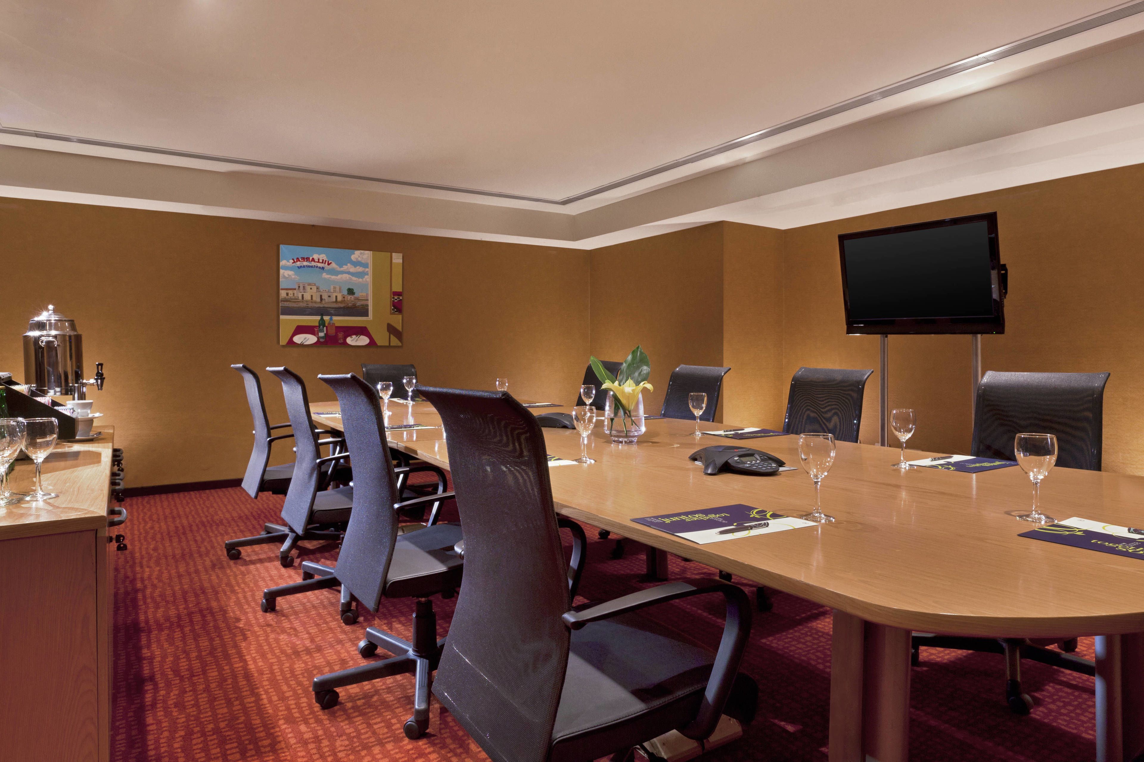 A conference room with a long table, six chairs, a TV screen, and a poster on the wall.