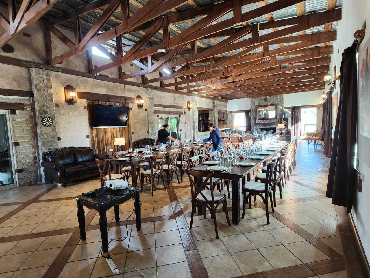 A spacious dining area with high ceilings and exposed beams, featuring multiple tables and chairs, a stone wall, and a television mounted on the wall.