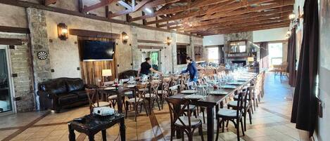A spacious dining area with high ceilings and exposed beams, featuring multiple tables and chairs, a stone wall, and a television mounted on the wall.