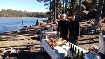 An outdoor dining setup with a table set for a meal, surrounded by natural scenery and a body of water.