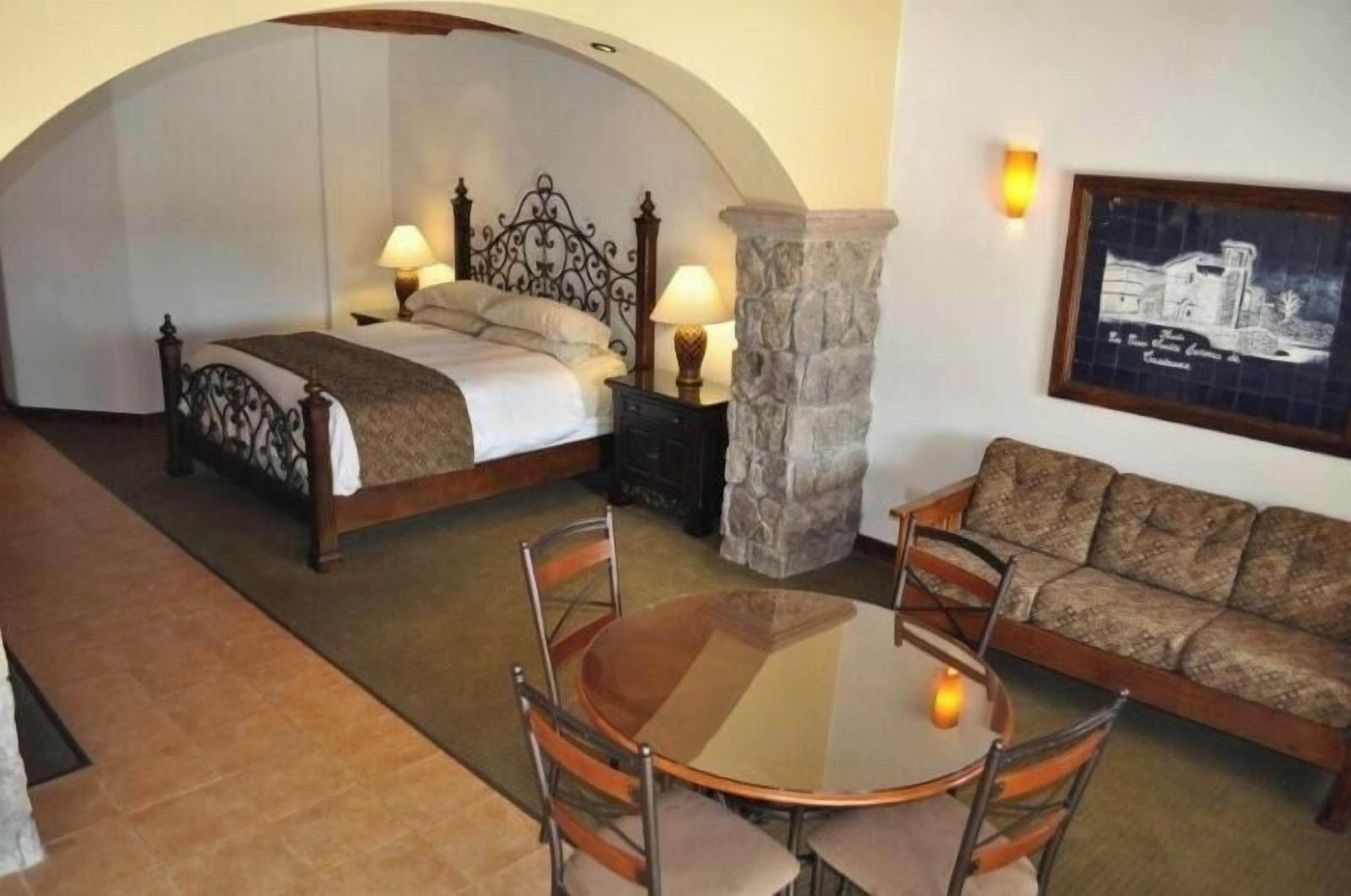 A bedroom with a bed, bedside tables, a stone pillar, a sofa, and a table with chairs.