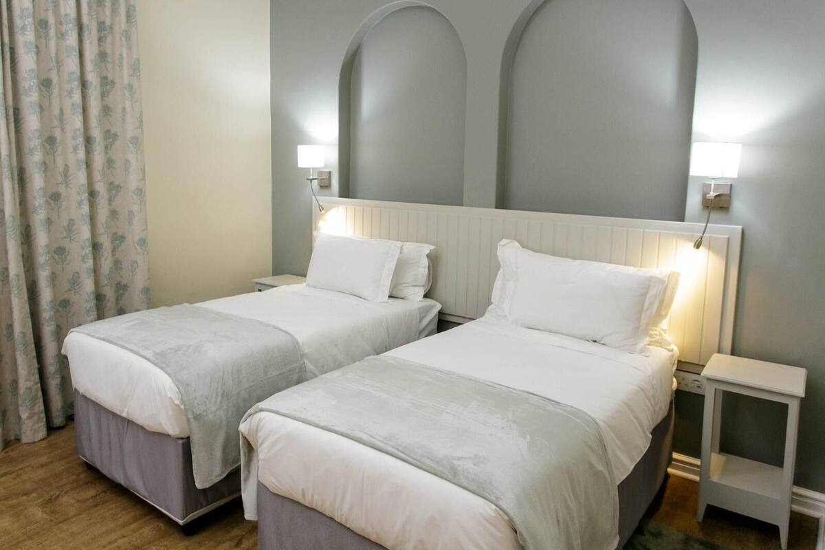 A hotel room with two single beds, each with a white bedspread and matching pillows. There are bedside tables with lamps, and the room has a simple, clean design with a light-colored wall and a wooden floor.