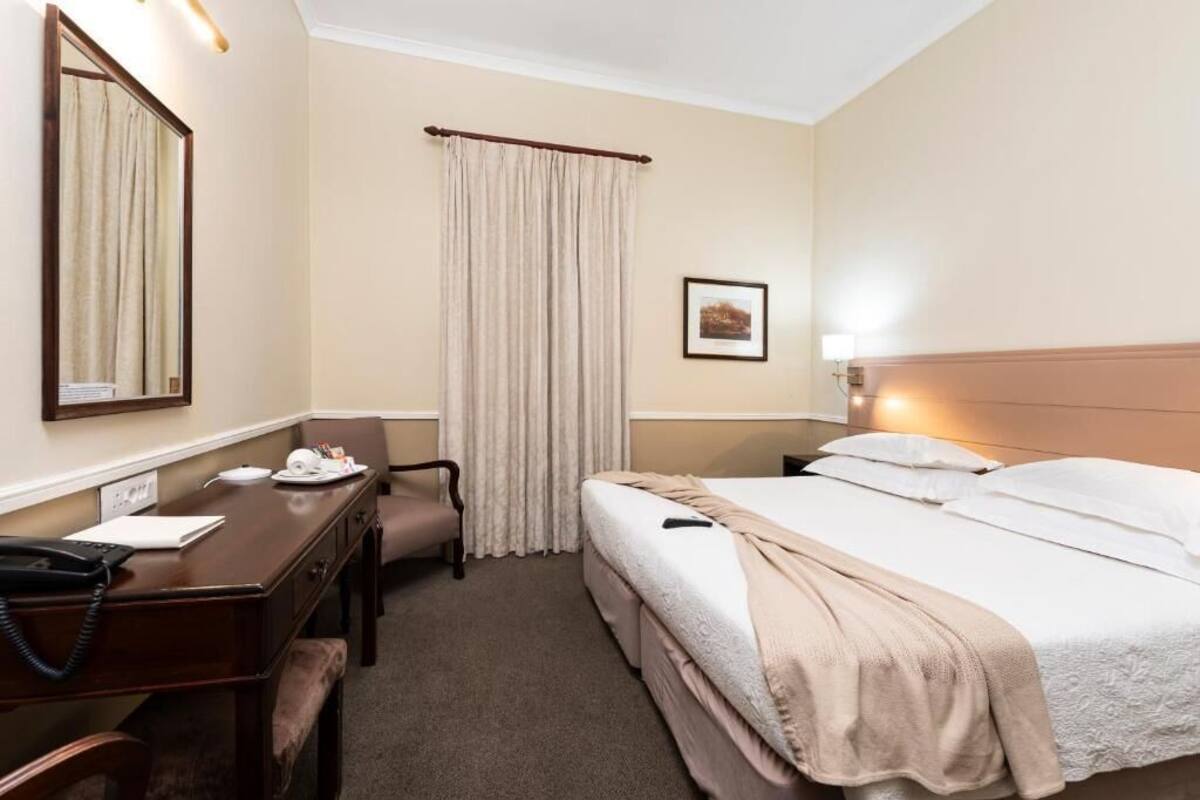 A hotel room with a bed, a desk with a telephone, a chair, a window with curtains, and a framed picture on the wall.