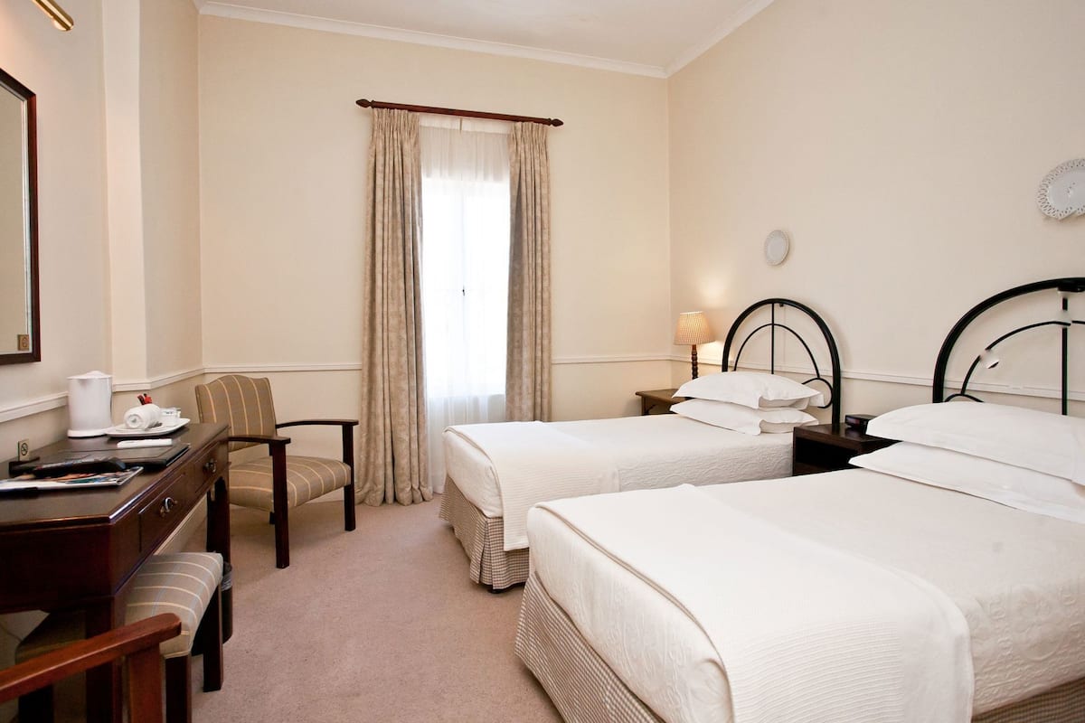 A hotel room with two beds, a desk with a chair, a lamp, and a window with curtains.