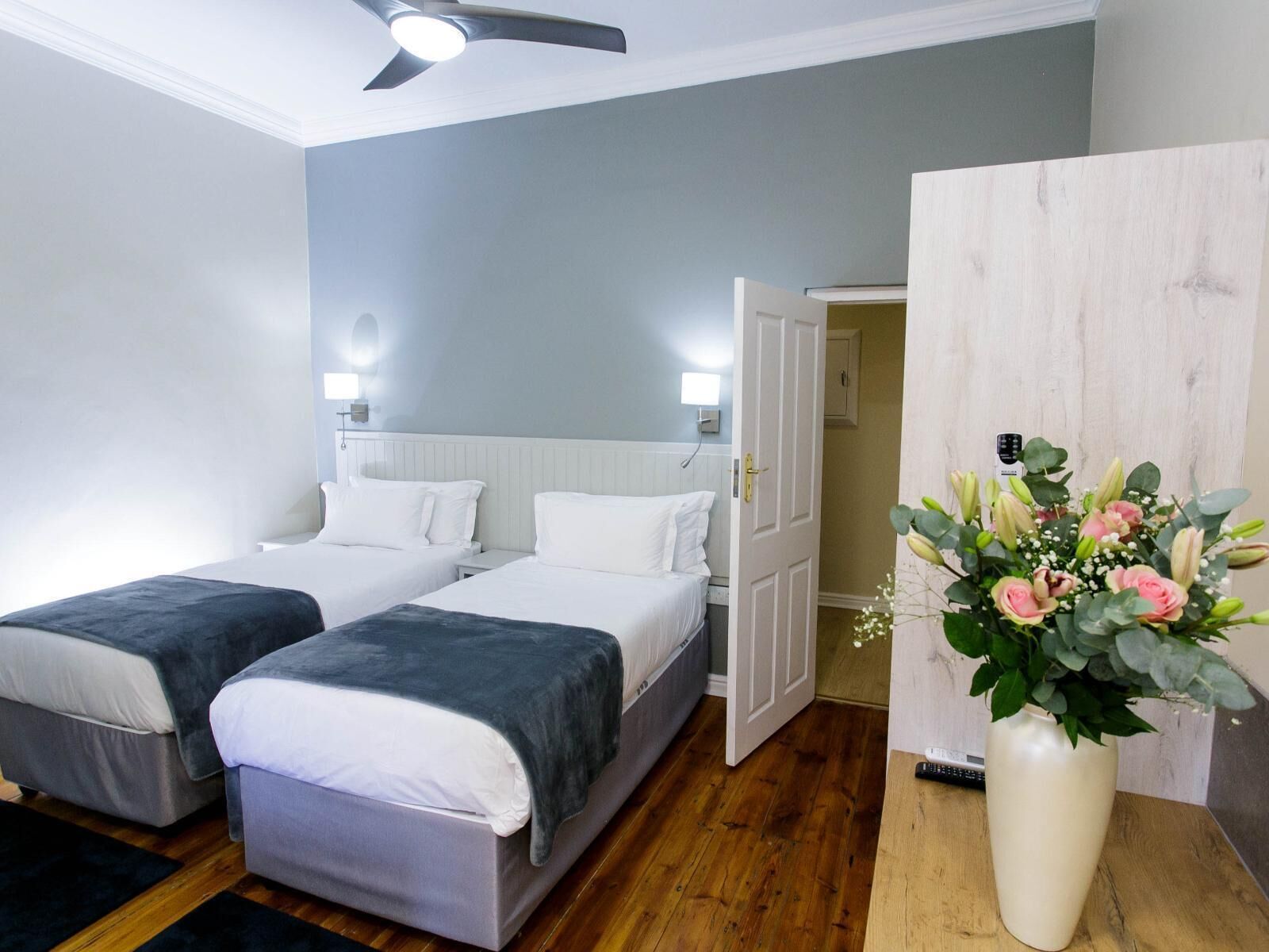 Una habitación de hotel con dos camas, un ventilador de techo, una mesita de noche con lámpara y un jarrón con flores.
