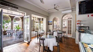 Un interior de cafetería con mesas redondas, sillas, un panel con la carta y una vitrina con pasteles.
