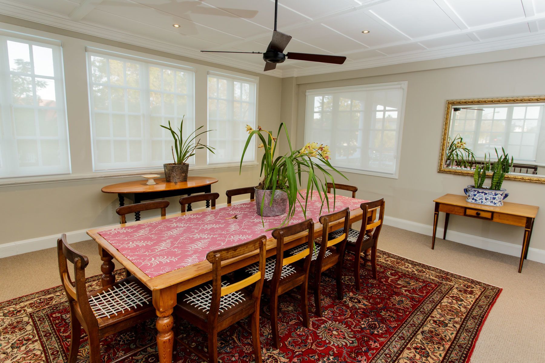 A dining room with a wooden table, six chairs, and a ceiling fan. There are potted plants on the table and a rug on the floor.