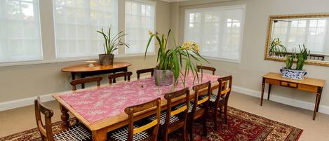 A dining room with a wooden table, six chairs, and a ceiling fan. There are potted plants on the table and a rug on the floor.