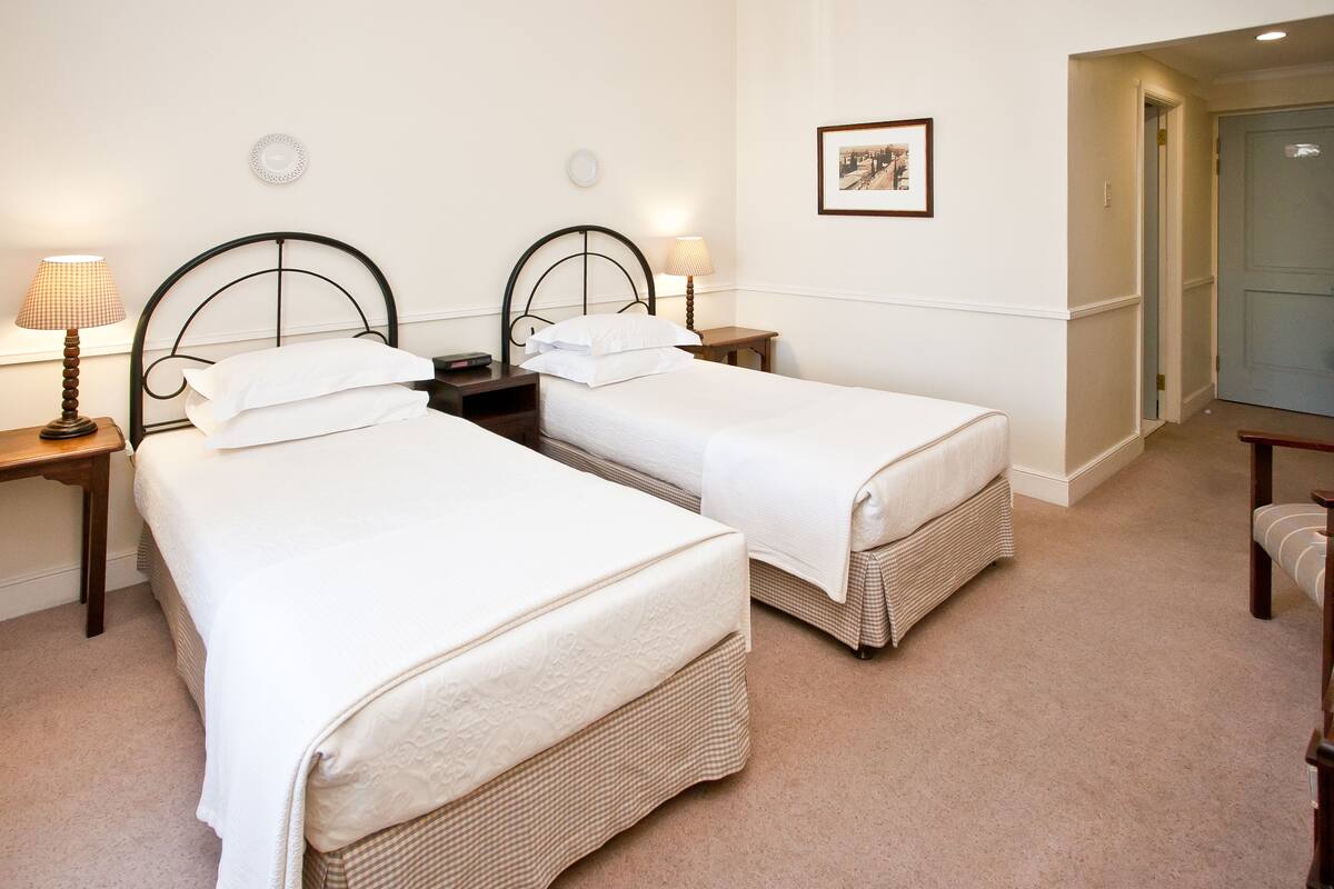 A hotel room with two single beds, a small table, two bedside lamps, a framed picture on the wall, and a door leading to another room.