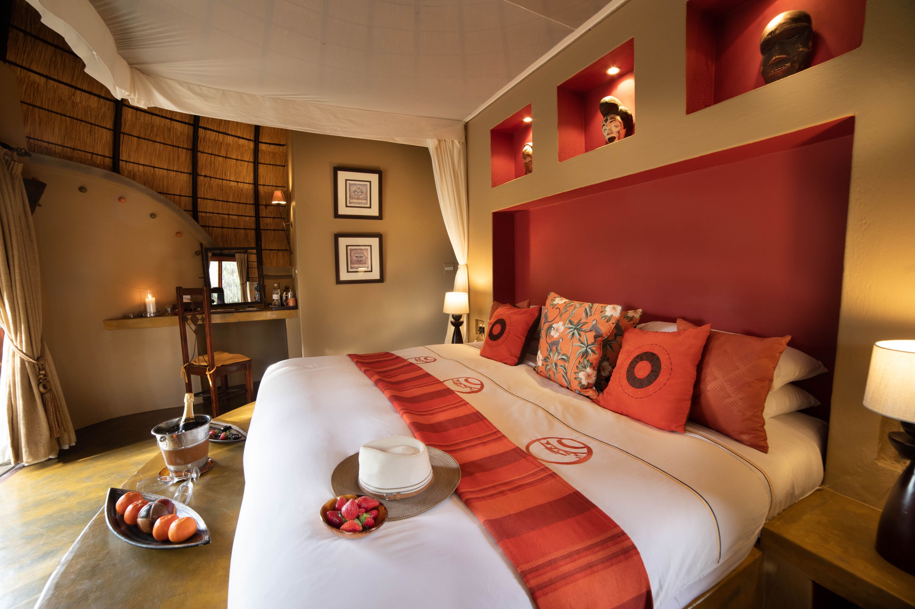 Quarto de hotel com uma cama grande, uma mesa de jantar com frutas e uma bebida, e vista para o exterior através de uma janela.