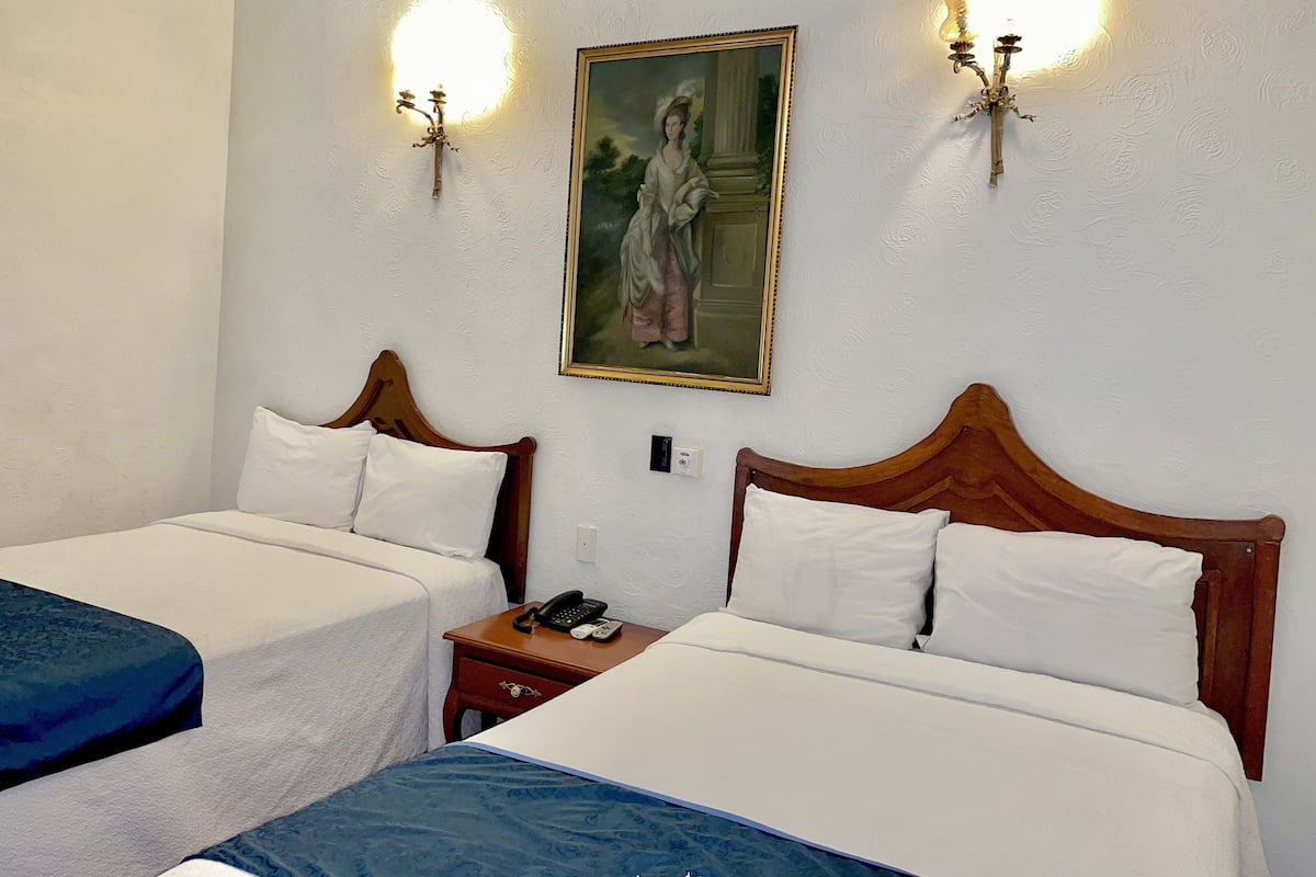 A hotel room with two beds, a painting on the wall, and two wall-mounted lamps.