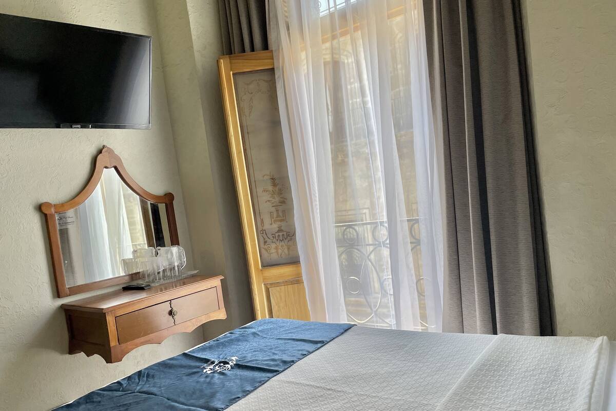 Una cama bien hecha con cubrecama azul, una mesita de noche de madera con un espejo y vasos, un televisor montado en la pared y una ventana con cortinas de gasa.