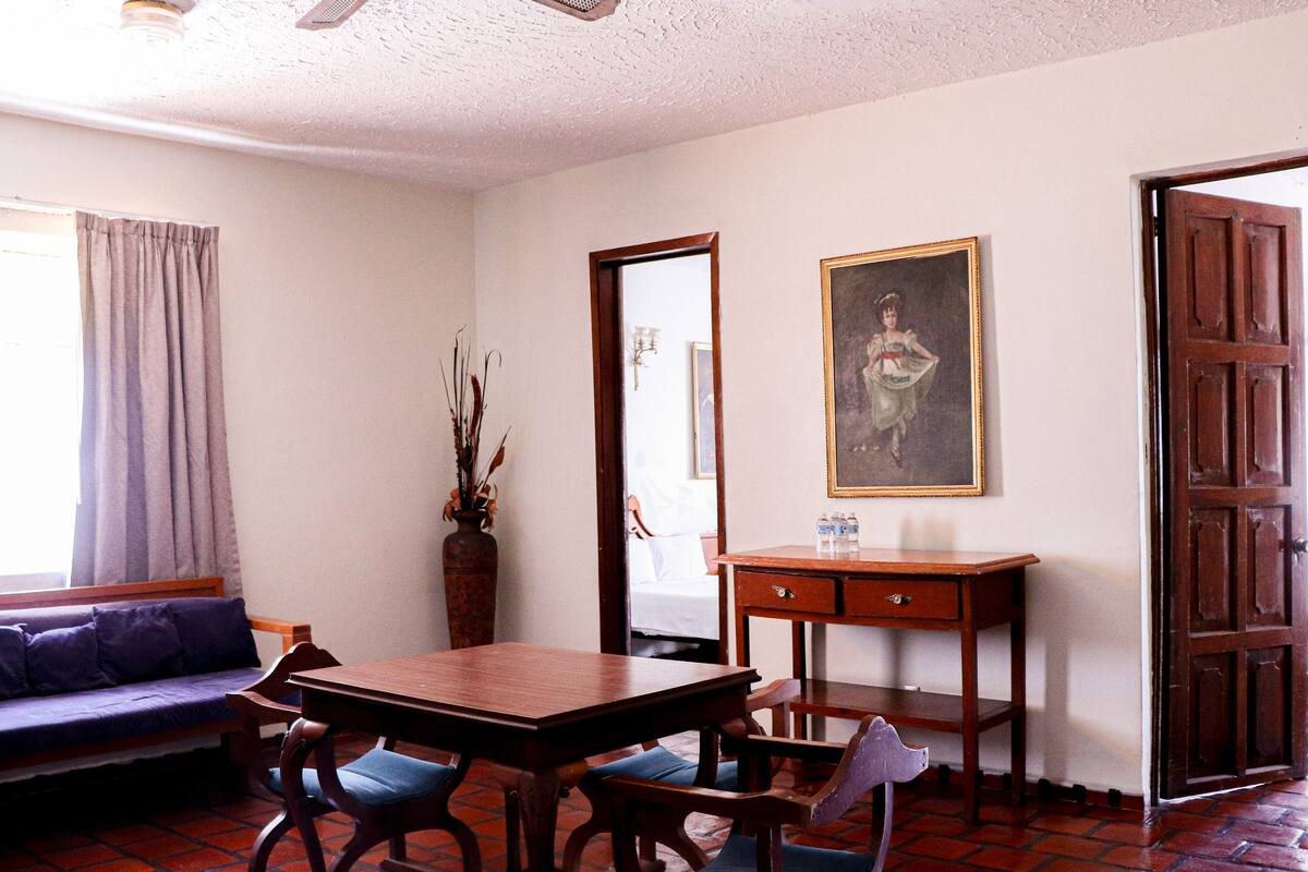 A room with a wooden table, chairs, a sofa, a vase with dried plants, a painting, and a wooden door.