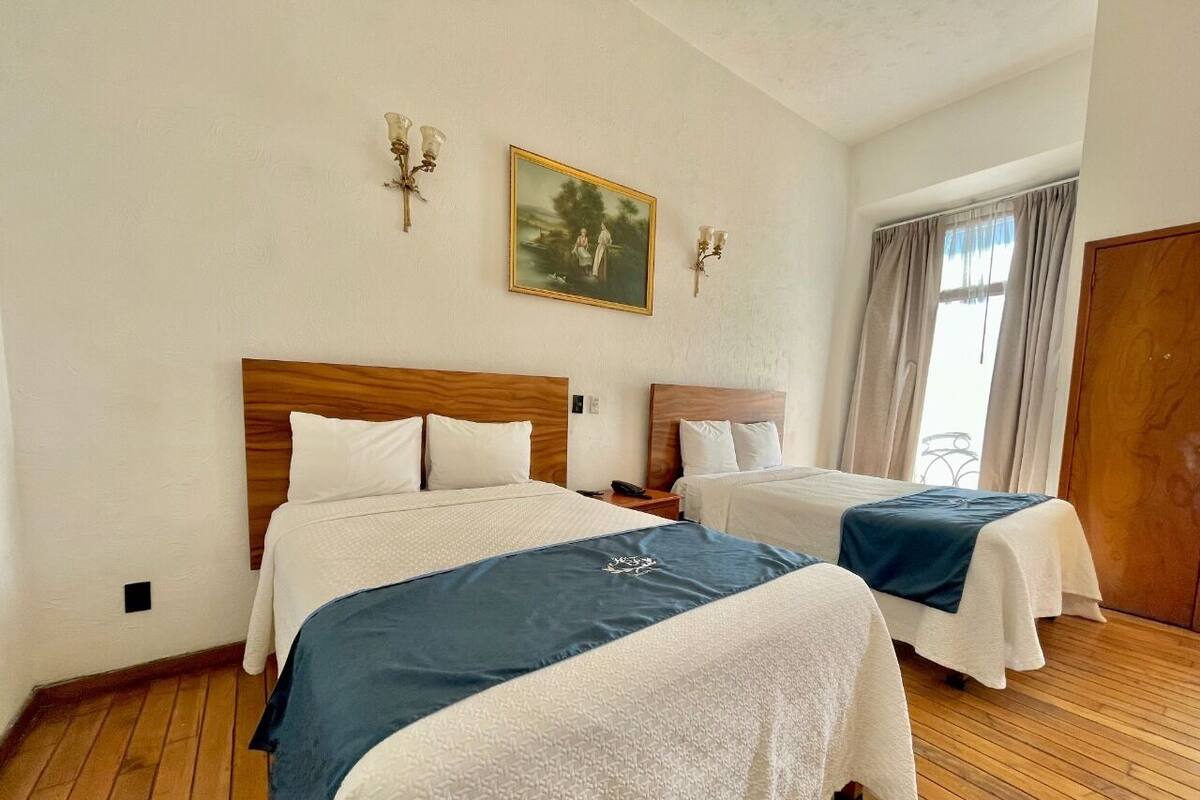 A hotel room with two beds, wooden headboards, white bedding, a painting on the wall, and a window with curtains.